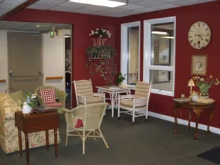 A cozy common area with red walls featuring a small seating arrangement including wicker chairs with cushions, a floral sofa, a small table with plants, a wall clock, and framed artwork. There are two windows and a door leading to another room.
