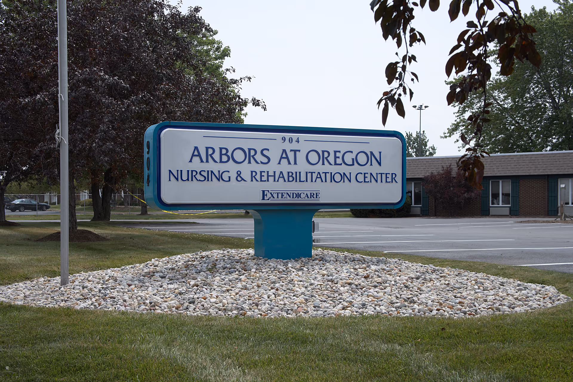 Outdoor view of a sign for Arbors at Oregon Nursing & Rehabilitation Center, mounted on a blue pedestal surrounded by white rocks, with trees and a parking lot in the background.