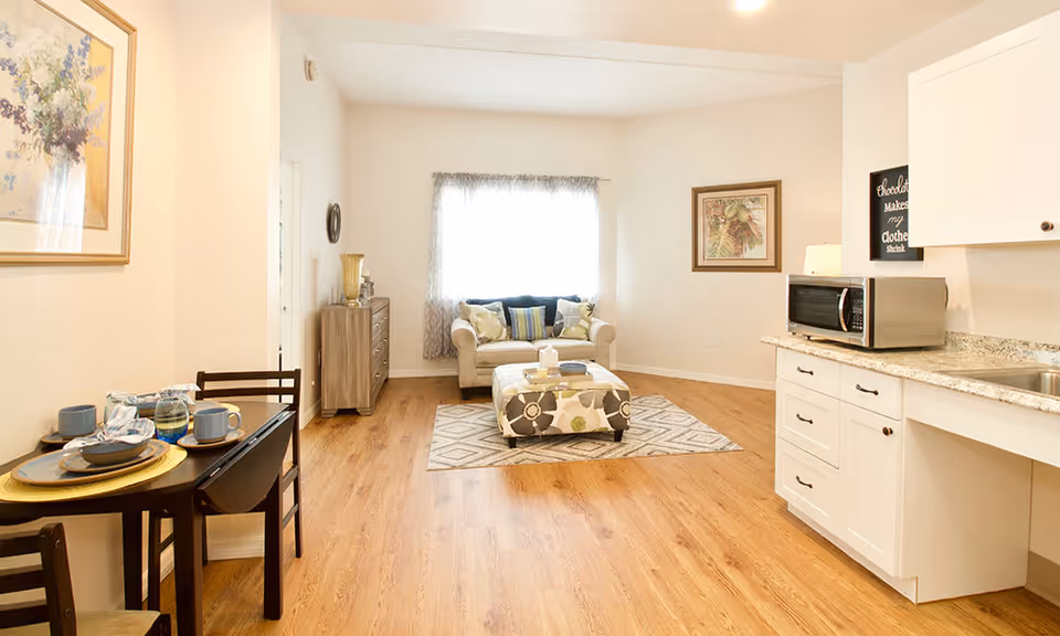 A bright and cozy living area with light wood flooring. There is a small dining table set with plates, cups, and napkins on the left. In the center, a beige loveseat with colorful cushions sits in front of a window with sheer curtains. A patterned ottoman is placed on a geometric rug in front of the loveseat. On the right, a kitchen counter with white cabinets, a microwave, and a sink is visible. The walls are decorated with framed artwork.