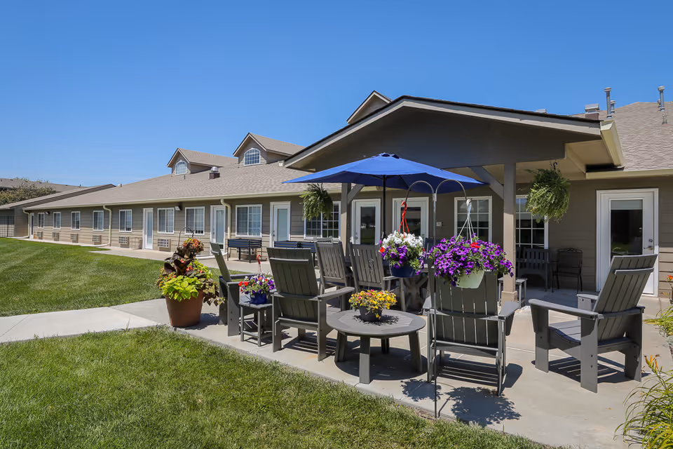 Outdoor patio area at Homestead Assisted Living of La Vista featuring several gray wooden chairs and tables arranged under a covered structure with a blue umbrella. There are hanging flower pots with purple and white flowers, potted plants, and a well-maintained green lawn surrounding the patio. The building exterior is beige with multiple windows and doors under a clear blue sky.