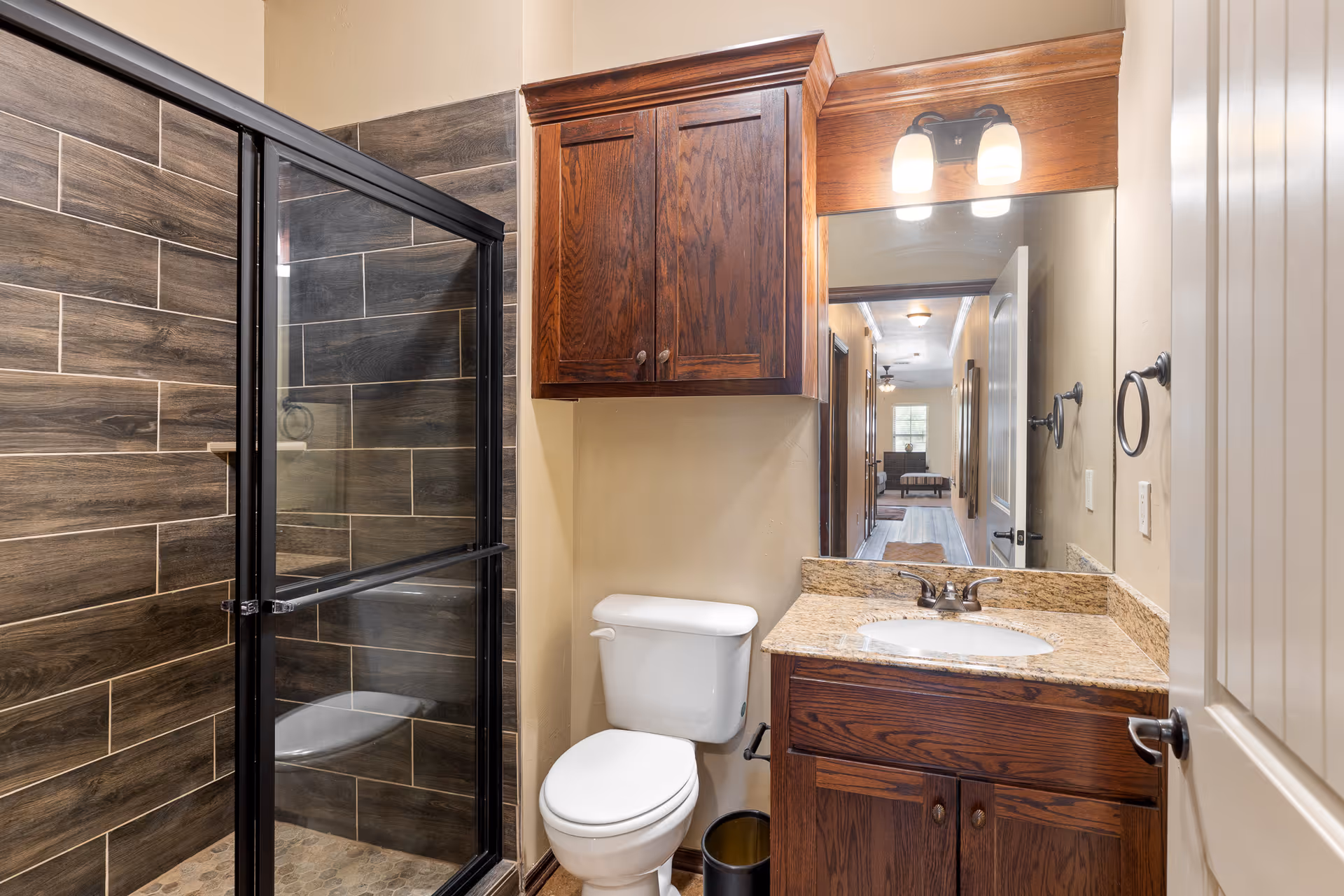 Bathroom with a glass-enclosed shower featuring dark wood-look tile, a white toilet, and a wooden vanity with granite countertop and mirror.