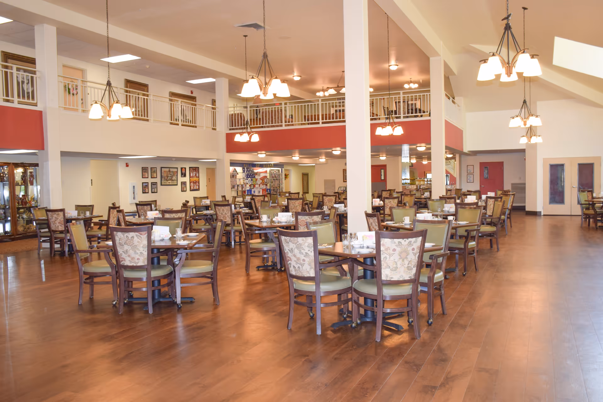 Spacious dining room with multiple wooden tables and chairs arranged neatly on a polished wooden floor. The room features high ceilings with hanging light fixtures and a second-floor balcony railing overlooking the area. Walls are decorated with framed pictures and there are large double doors on the right side.