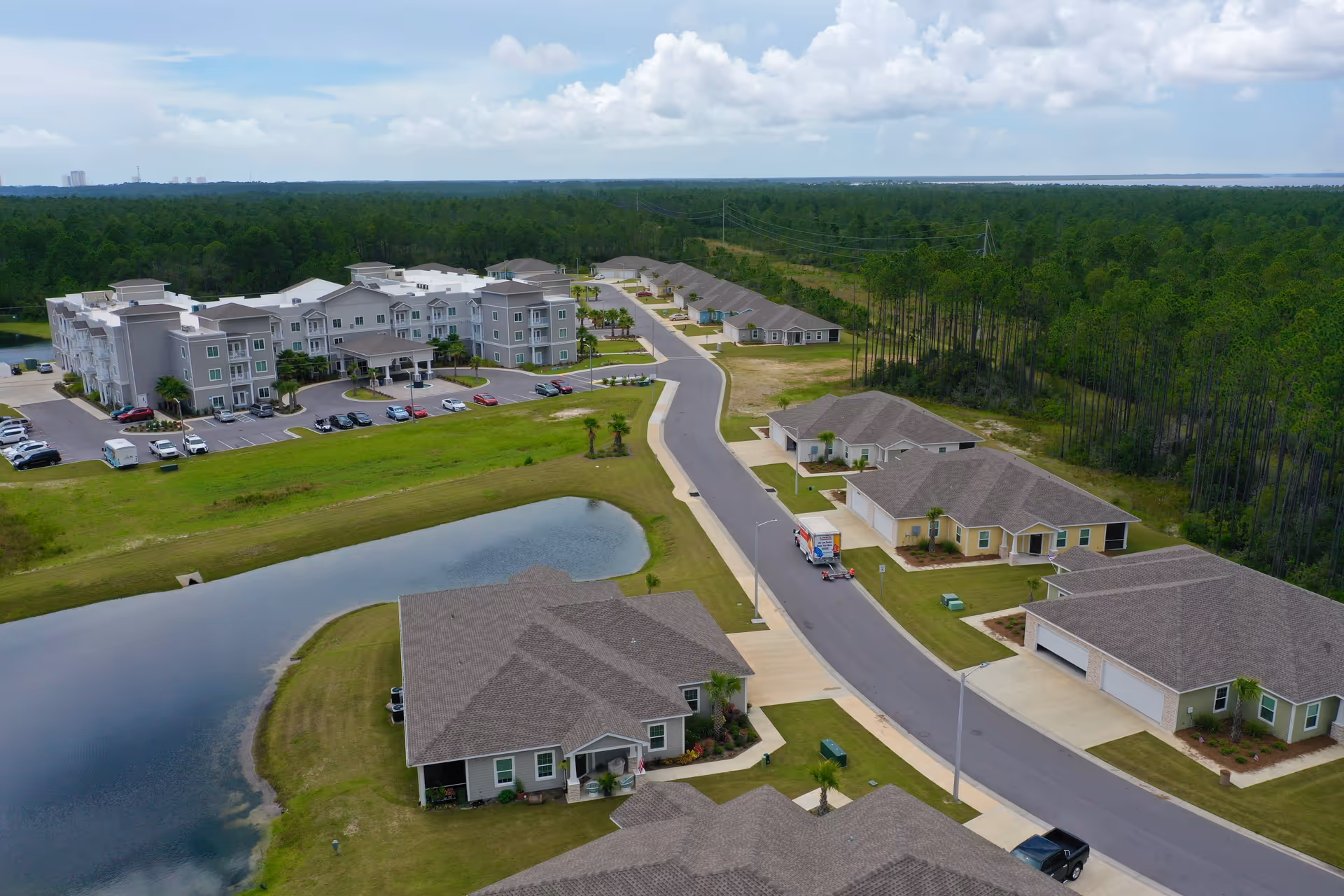 Aerial view of Seagrass Village Independent Living facility showing multiple residential buildings, a pond, a road with parked vehicles, and surrounding green forested area under a partly cloudy sky.