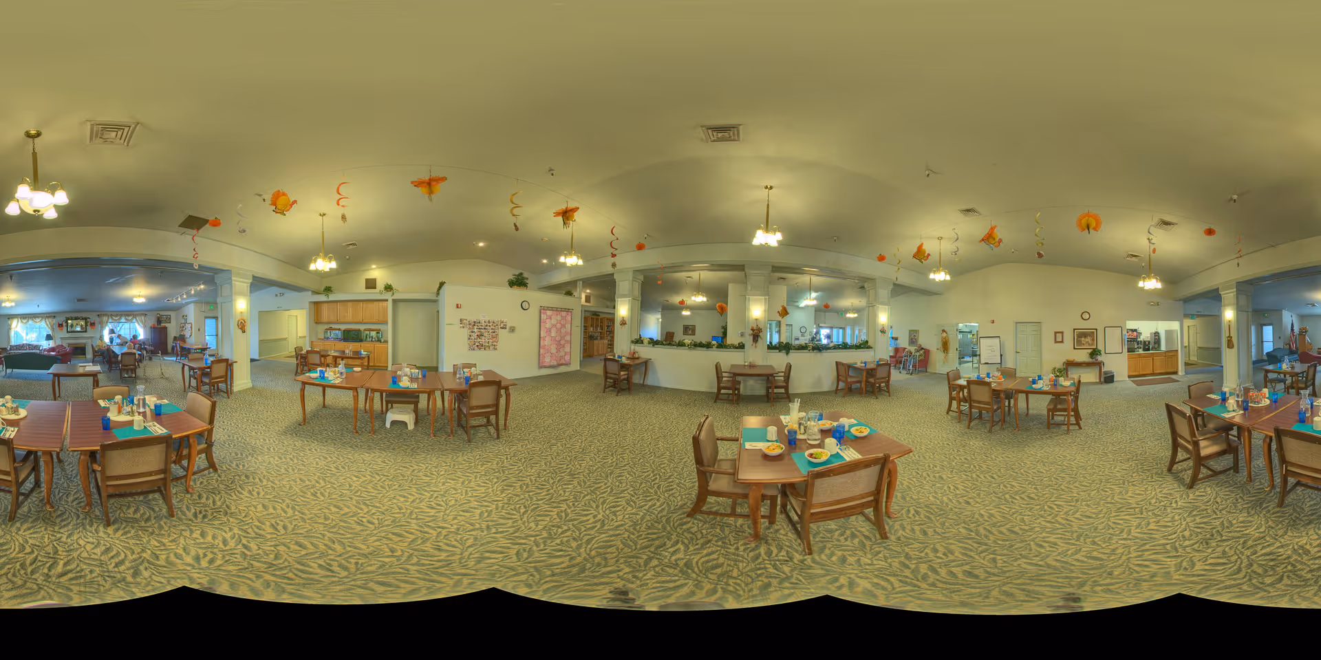 A spacious dining room in an assisted living facility with multiple wooden tables and chairs arranged neatly. Tables are set with plates, cups, and utensils, ready for a meal. The room has carpeted flooring with a leafy pattern, white walls, and several ceiling lights. Decorative orange and yellow hanging ornaments are suspended from the ceiling. There is a serving counter area and a kitchen visible in the background.