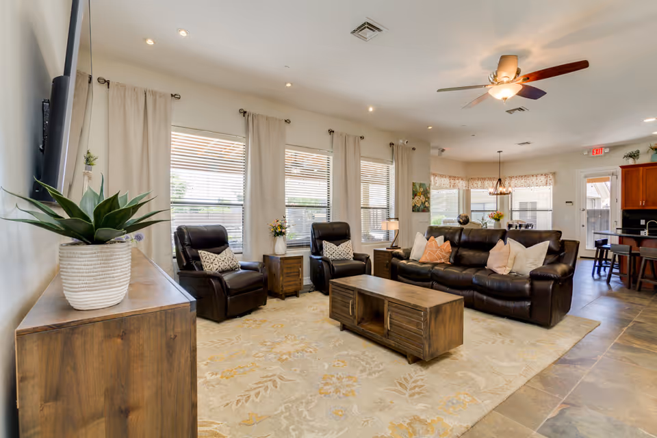 A bright and spacious living room with large windows covered by light curtains. The room features two dark leather recliners and a matching leather sofa adorned with decorative pillows. A wooden coffee table sits on a patterned area rug, and a wooden cabinet with a potted plant is visible in the foreground. The ceiling has recessed lighting and a ceiling fan with a light fixture. In the background, there is a dining area and kitchen with wooden cabinetry.