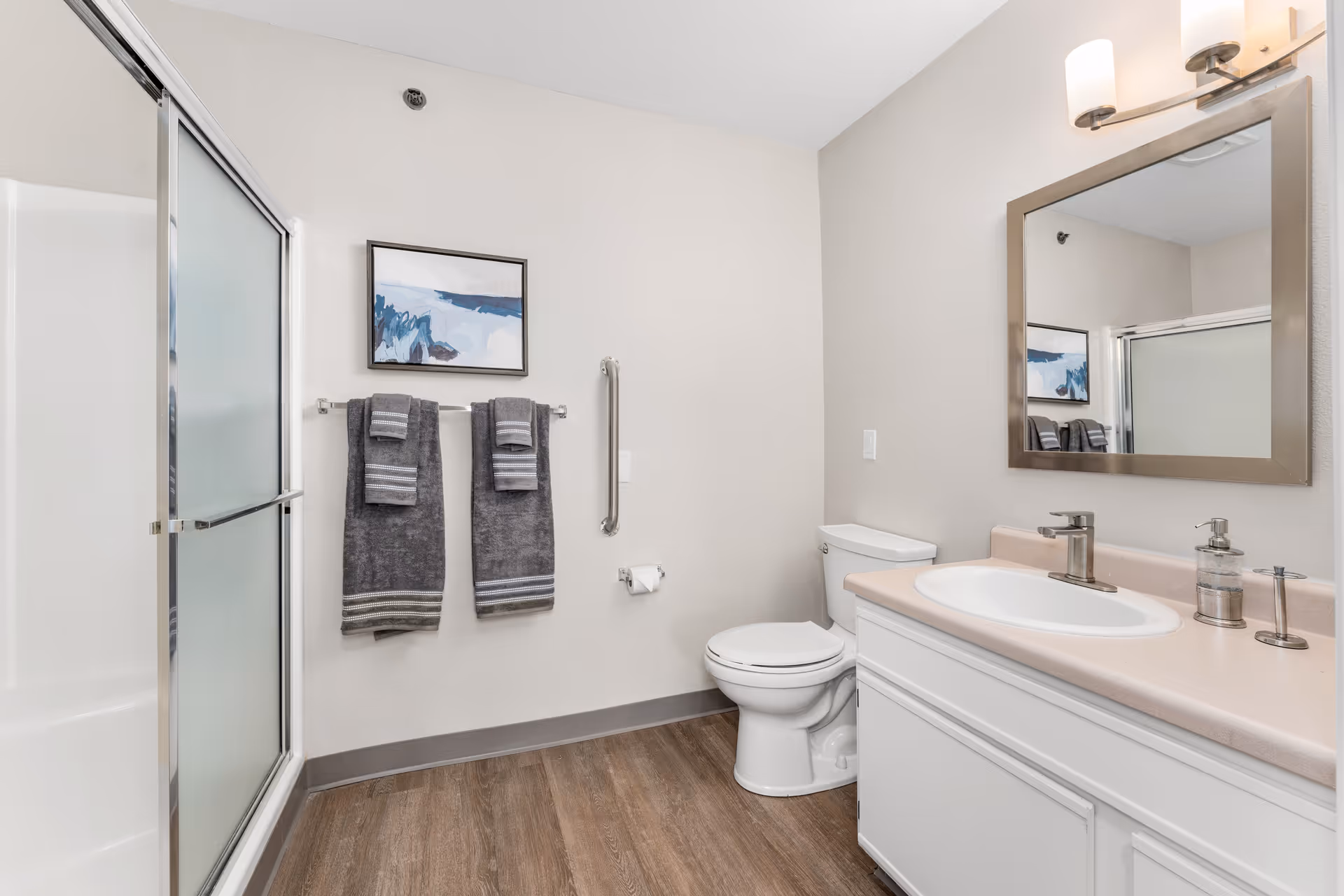 A clean and modern bathroom featuring a white toilet, a white vanity with a sink and a large mirror above it, a shower with sliding frosted glass doors, two gray towels hanging on a towel rack, a grab bar on the wall, and a framed abstract blue and white painting. The floor has wood-like vinyl flooring and the walls are painted light beige.