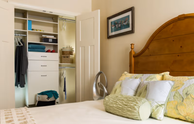 Sunlit bedroom with a wooden headboard, decorative pillows on the bed, and an open closet showing shelving and hanging clothes.