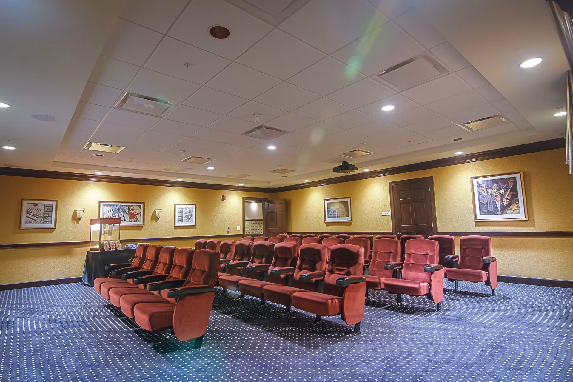 Interior view of a small theater or screening room with rows of red cushioned seats arranged on a blue carpet with white dots. The walls are yellow with framed artwork, and there is a popcorn machine on a table in the back corner. The ceiling has recessed lighting and air vents.