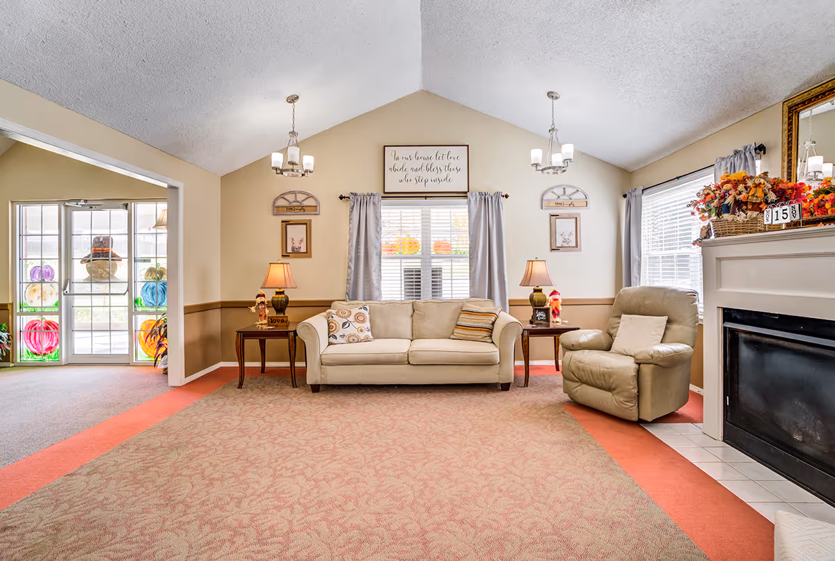 A cozy living room with a beige sofa and a light brown recliner chair. Two side tables with lamps flank the sofa. The walls are decorated with framed pictures and a sign above the window that reads 'In our home let love abide and bless those who step inside.' There is a fireplace on the right side adorned with autumn-themed decorations. The room has a high vaulted ceiling and carpeted floor with a red border. Through an open doorway on the left, a glass door with colorful pumpkin decorations is visible.