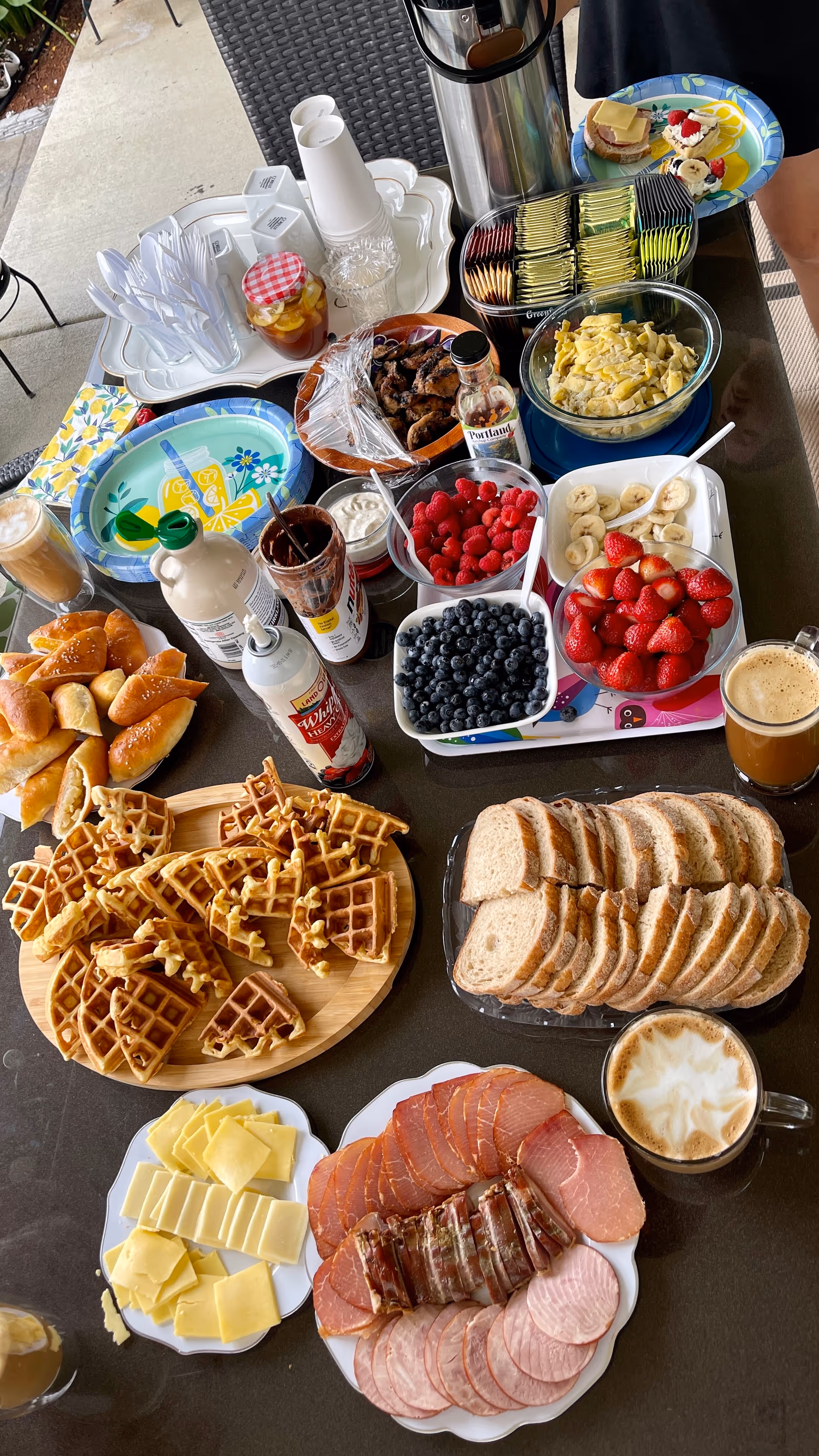 A table set with a variety of breakfast foods including waffles, sliced bread, assorted cheeses, cold cuts, fresh berries (blueberries, raspberries, strawberries, banana slices), croissants, a bowl of scrambled eggs, condiments, coffee cups, and a tray with plastic utensils and cups.
