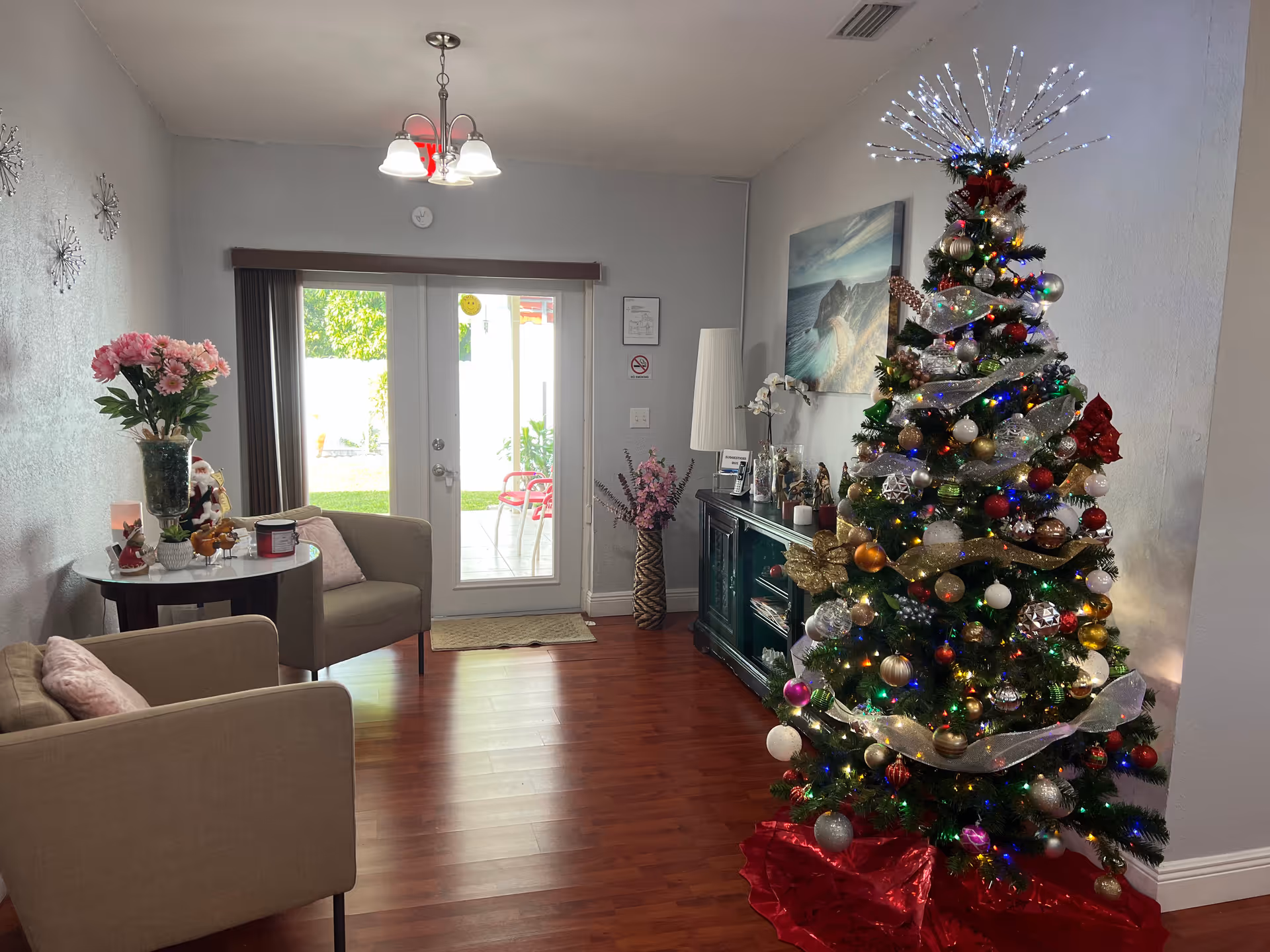 A cozy living room decorated for Christmas with a large, decorated Christmas tree on the right side. The tree is adorned with colorful lights, ornaments, and a starburst light topper. There are two beige armchairs with pink cushions on the left side, a small round table with flowers and holiday decorations between them. A green cabinet with decorative items and a lamp is against the far wall. French doors with glass panes lead to an outdoor patio with red chairs and greenery visible outside. The room has wooden flooring and light gray walls.