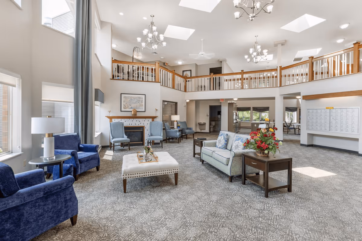 A spacious and well-lit senior living facility common area with high ceilings and skylights. The room features comfortable seating including blue armchairs, a light blue sofa, and a white cushioned ottoman. There is a fireplace with a framed picture above it, side tables with lamps, and a table with a flower arrangement. A balcony railing overlooks the area, and there are mailboxes mounted on the wall to the right.