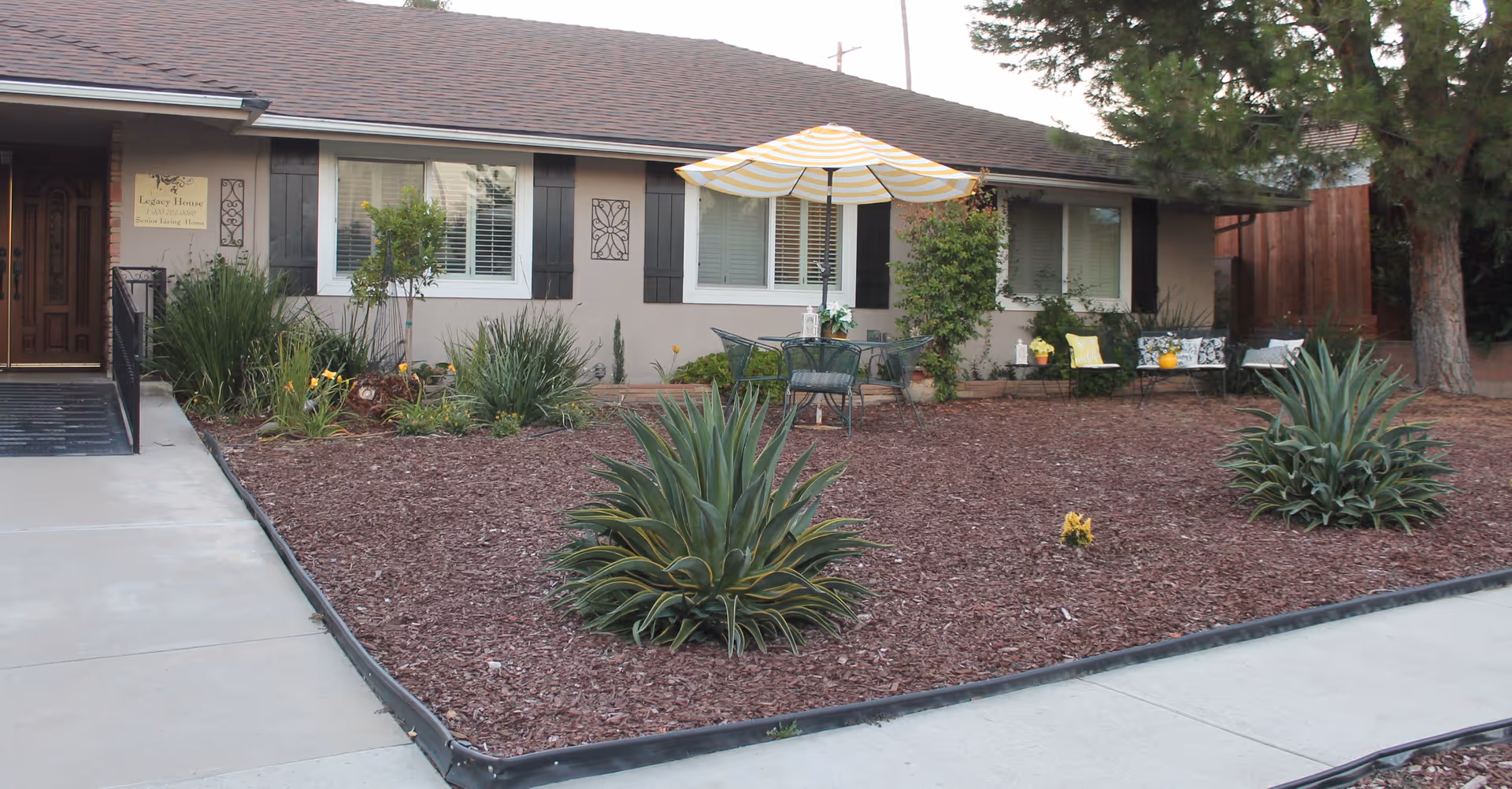 Front exterior view of Legacy House Upland, showing a single-story building with a brown roof, beige walls, and black window shutters. The landscaped front yard features mulch, green plants, a patio table with a yellow and white striped umbrella, and outdoor seating with cushions. A concrete walkway leads to the entrance with a ramp and handrails.