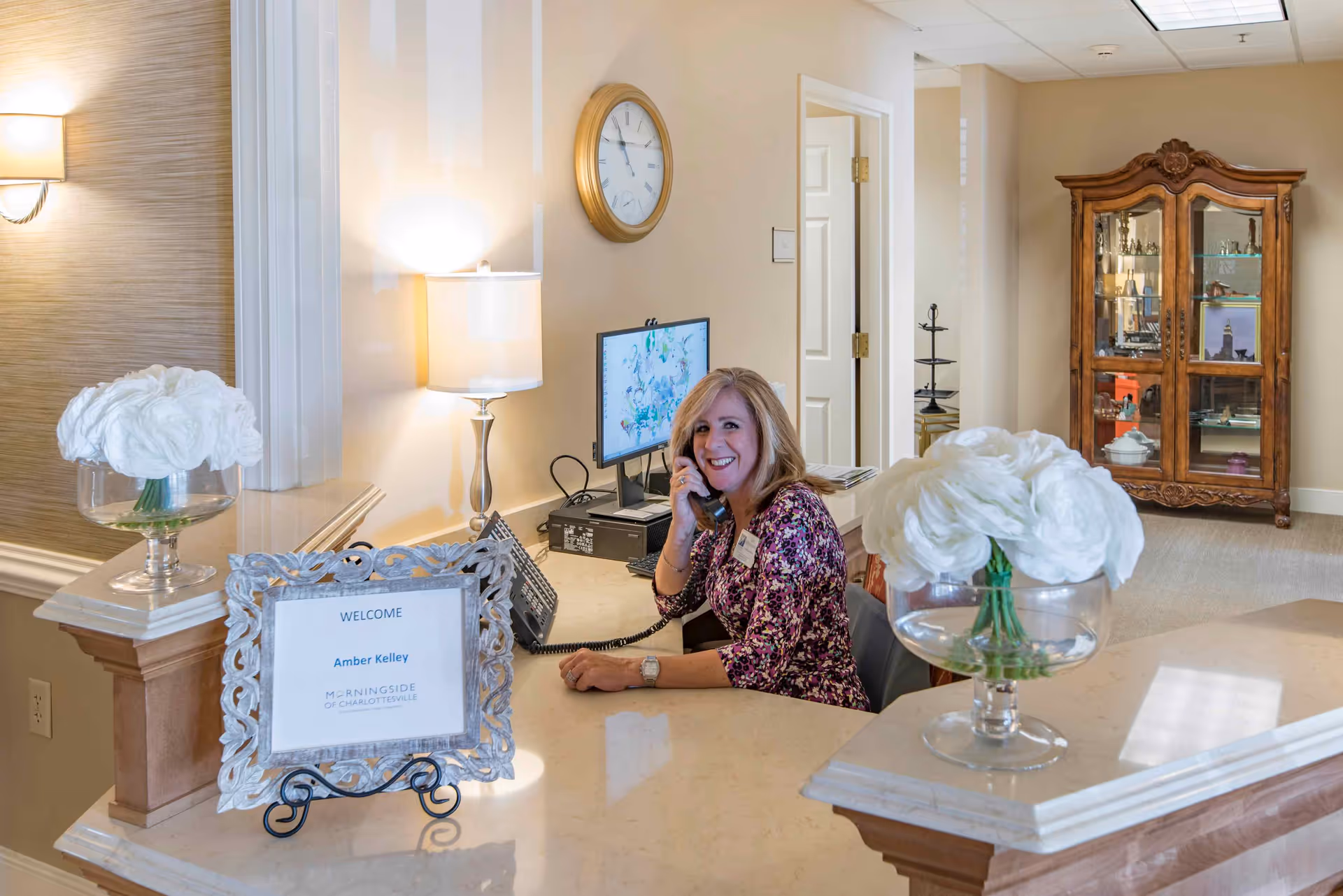 Reception area at Morningside of Charlottesville with a smiling woman sitting behind the desk, talking on the phone. The desk has two glass vases with white flowers and a framed welcome sign for Amber Kelley. A computer monitor and a clock on the wall are visible in the background, along with a wooden display cabinet.