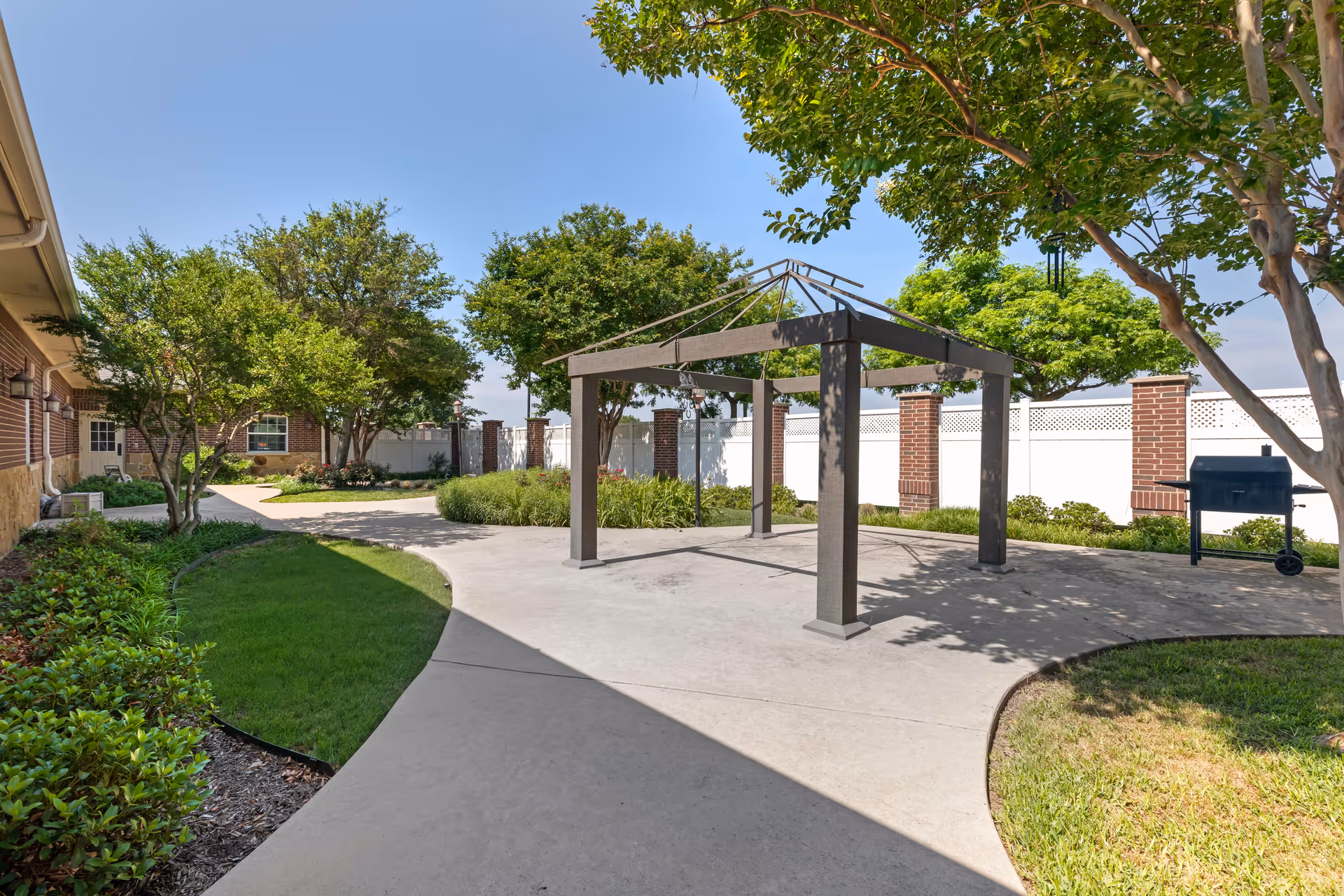 Outdoor courtyard area with a concrete pathway, green grass, and trees. There is a metal gazebo frame without a canopy in the center and a black barbecue grill to the right. The area is enclosed by a white fence with brick pillars, and part of a brick building is visible on the left side.