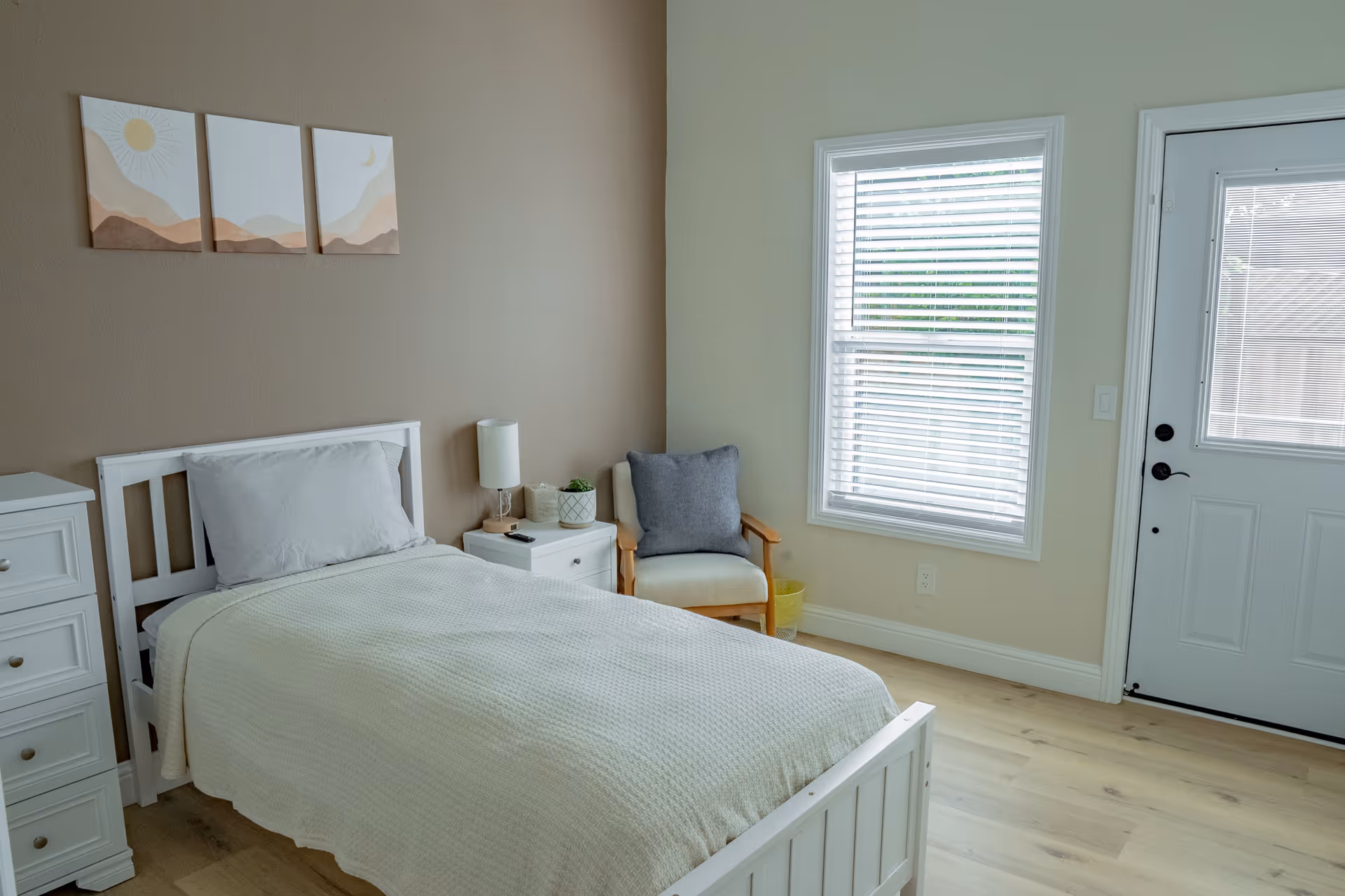 A simple, clean bedroom with a single white bed covered with a beige blanket and a gray pillow. Next to the bed is a white nightstand with a lamp, a small plant, and a remote control. There is a wooden armchair with a gray cushion near a window with white blinds. The walls are painted in neutral tones, and three minimalist art pieces hang above the bed. A white door is visible next to the window.