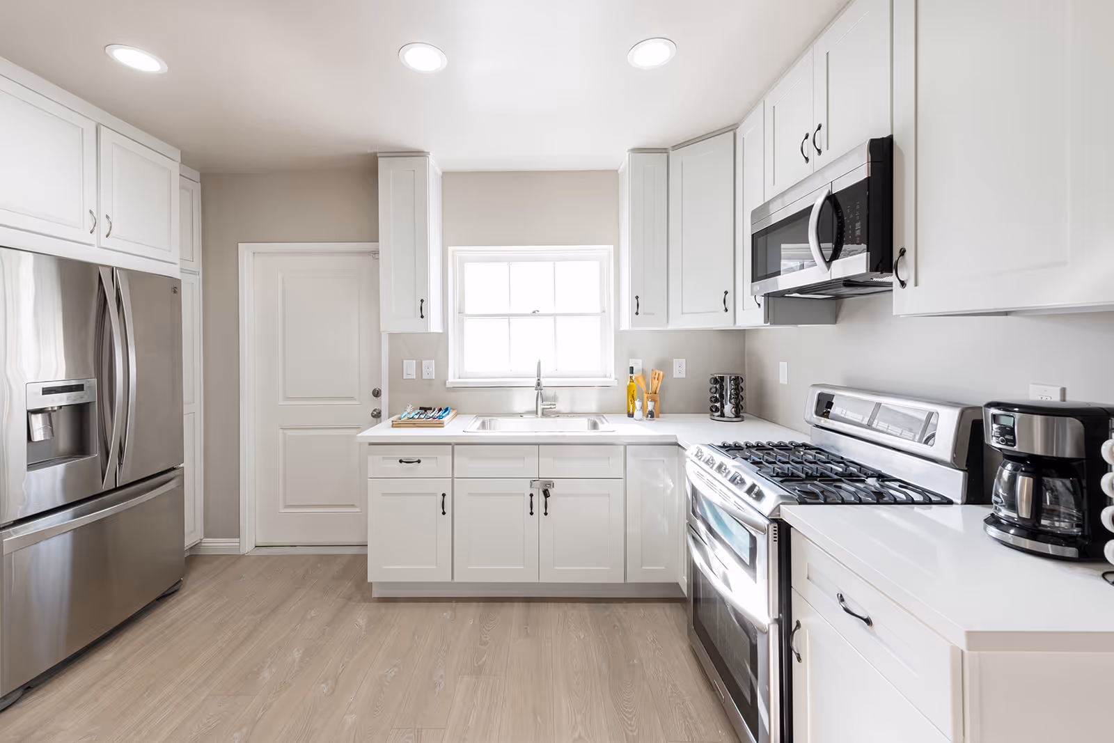 Bright and modern kitchen with white cabinets, stainless steel refrigerator, gas stove, microwave, coffee maker, and a window above the sink letting in natural light.