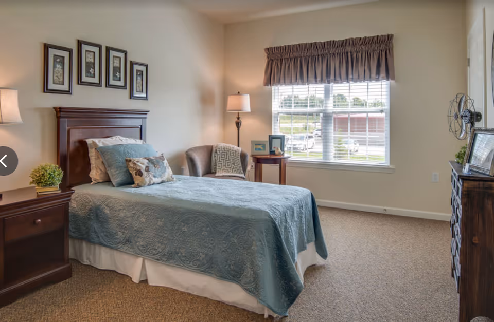 A neatly arranged bedroom with a single bed covered in a blue quilt and decorative pillows. There is a wooden nightstand with a lamp and a small plant on the left side of the bed. A comfortable armchair with a patterned throw blanket is placed near a window with blinds and a brown valance. A wooden dresser with framed pictures and a decorative fan is on the right side of the room. The walls are light-colored and decorated with four framed floral artworks above the bed.