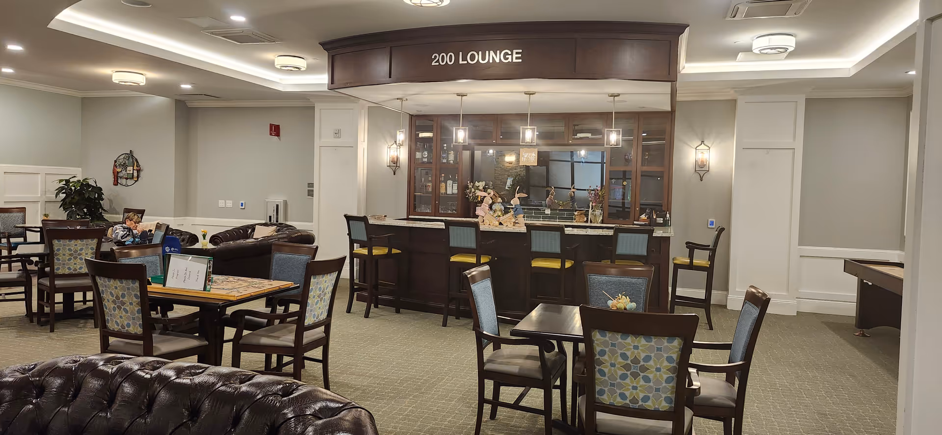 Interior view of a lounge area labeled '200 LOUNGE' with a bar counter and several bar stools. The room has multiple tables and chairs with patterned upholstery, a few leather sofas, and a person sitting on one of the sofas. The space is well-lit with ceiling lights and wall sconces, and there are decorative items on the bar counter.