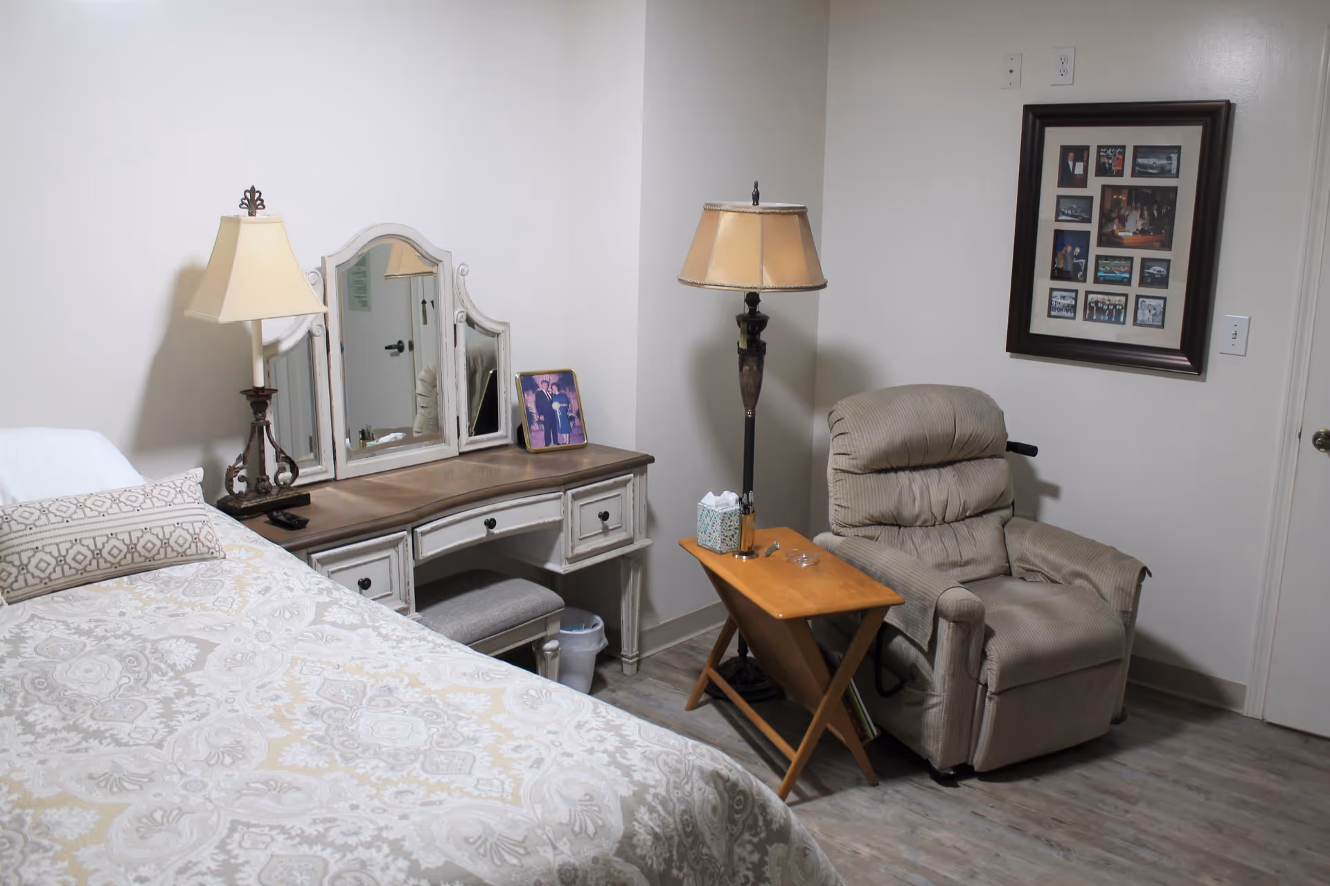 A cozy bedroom with a bed covered in a patterned beige and white bedspread, a wooden vanity with a tri-fold mirror and a lamp, a framed photo on the vanity, a beige recliner chair next to a wooden side table with a tissue box and a floor lamp behind it, and a framed collage of photos on the wall.