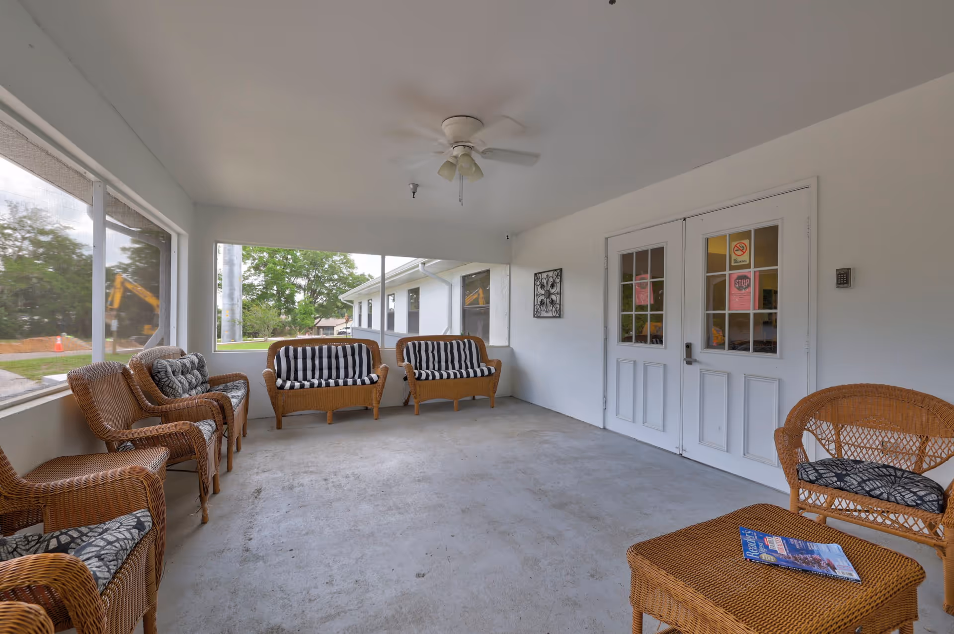 A covered outdoor seating area with wicker chairs and loveseats featuring patterned cushions. The space has a concrete floor, white walls, a ceiling fan, and large screened windows overlooking greenery. Double white doors with glass panes lead inside the building.