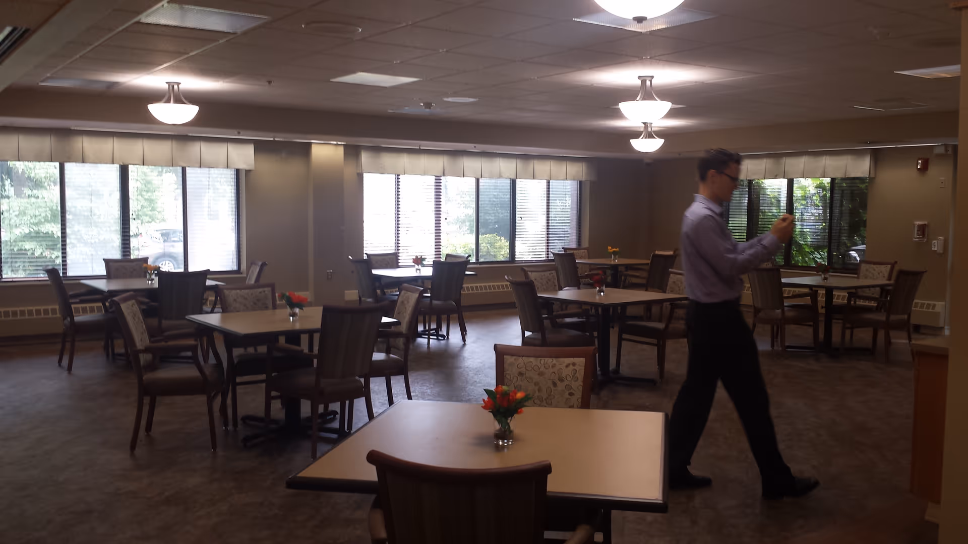 A dining room with multiple tables and chairs arranged neatly. Each table has a small vase with flowers. Large windows along the walls let in natural light. A man in a light purple shirt and dark pants is walking through the room, looking at his phone.