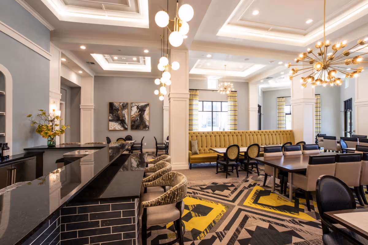 A spacious and elegant dining area with modern lighting fixtures, a long tufted yellow banquette, multiple tables with chairs, a black countertop bar with patterned bar stools, and abstract artwork on the walls. The room features high ceilings with recessed lighting and large windows with patterned curtains.