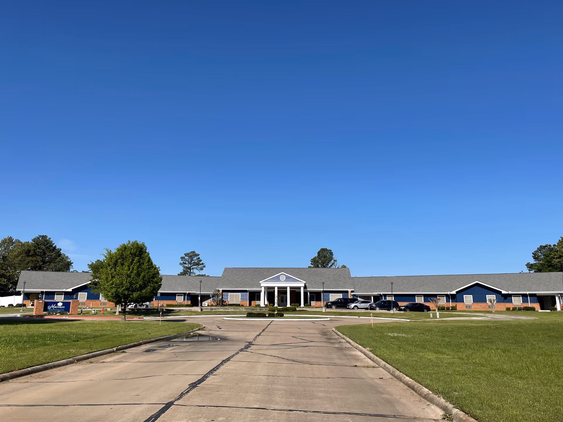 Front exterior view of Magnolia Assisted Living facility with a clear blue sky, a paved driveway leading to the entrance, green lawns on either side, and several parked cars near the building.
