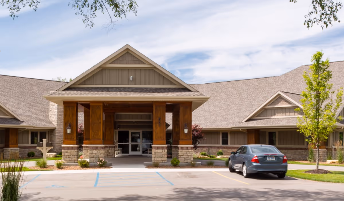 Front entrance of a single-story senior living facility with a covered porte-cochère, stone and wood columns and a parked car in the driveway.