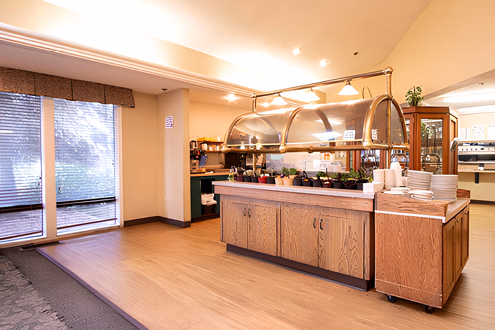 Interior view of a dining area with a wooden buffet counter featuring a glass sneeze guard and several potted plants on top. Plates and bowls are stacked on the counter. Large windows with blinds allow natural light to enter the room. The space has light-colored walls and wood flooring.