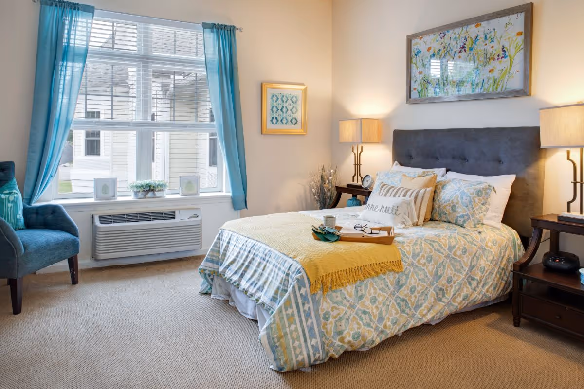 Sunlit bedroom with a made bed featuring patterned bedding and a yellow throw, bedside tables with lamps, and an armchair by a window with teal curtains.
