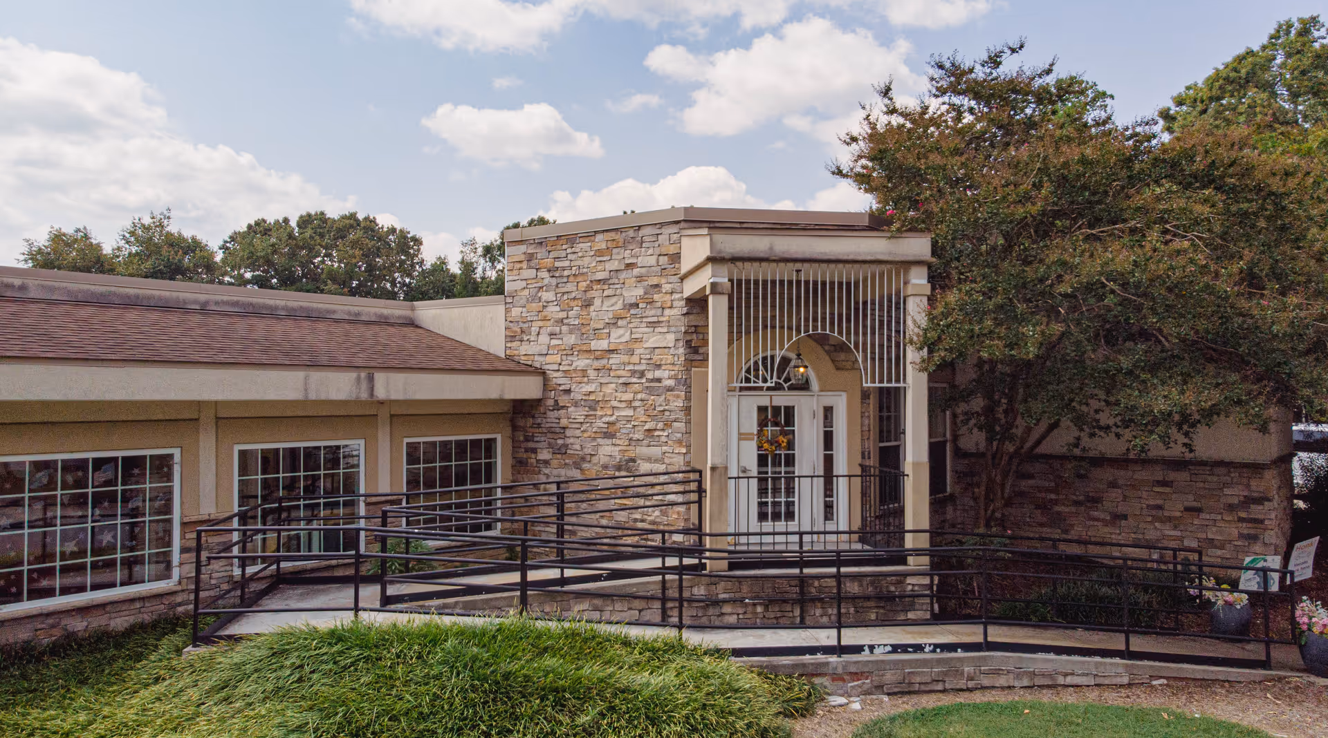 Exterior view of a building entrance with a stone facade and a white door with glass panels. There is a black metal ramp leading up to the door, surrounded by greenery and trees under a partly cloudy sky.