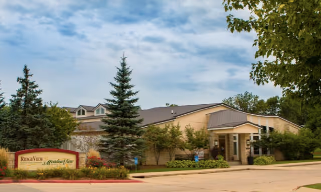 Exterior view of MeadowView Memory Care Village building with a sign in front that reads RidgeView and MeadowView. The building is surrounded by trees and landscaping under a partly cloudy sky.
