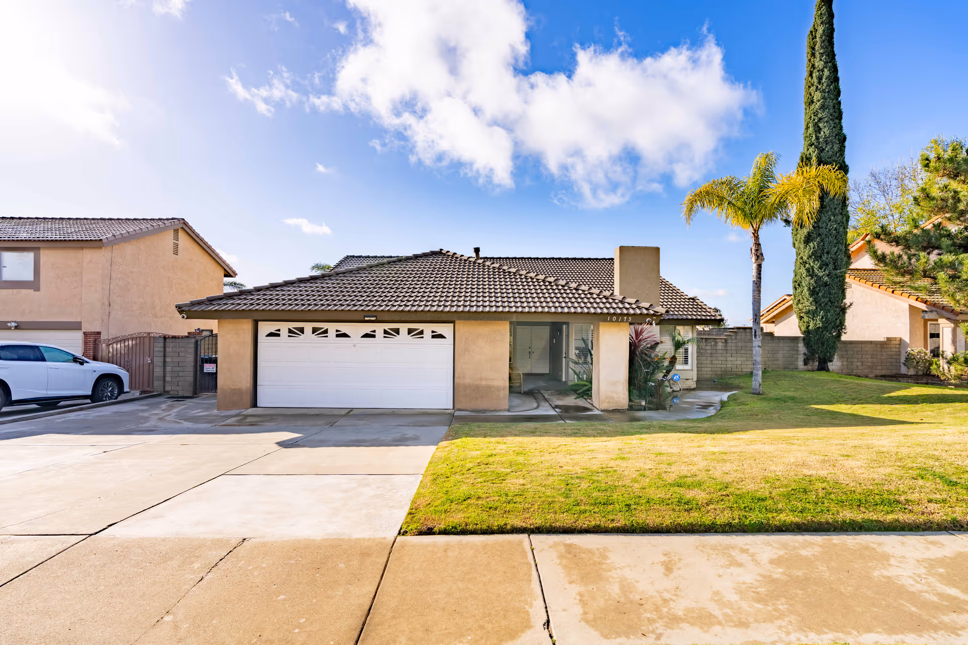 Front exterior view of a single-story house with a tiled roof, a double garage door, a driveway, a well-maintained lawn, and palm trees under a partly cloudy blue sky.