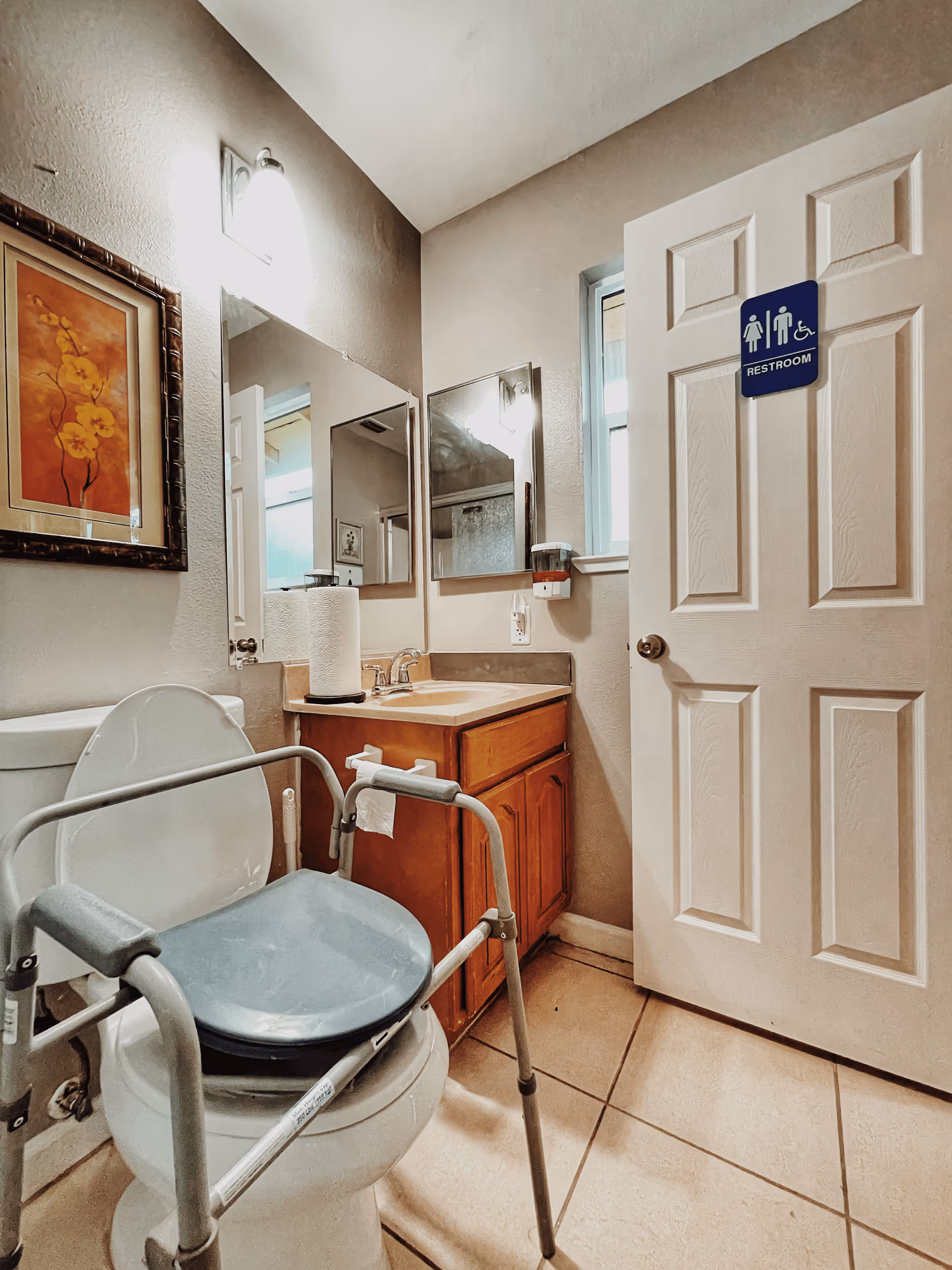 Small accessible bathroom with a raised commode frame over a toilet, a sink vanity with mirrors, and a door labeled 'Restroom'.
