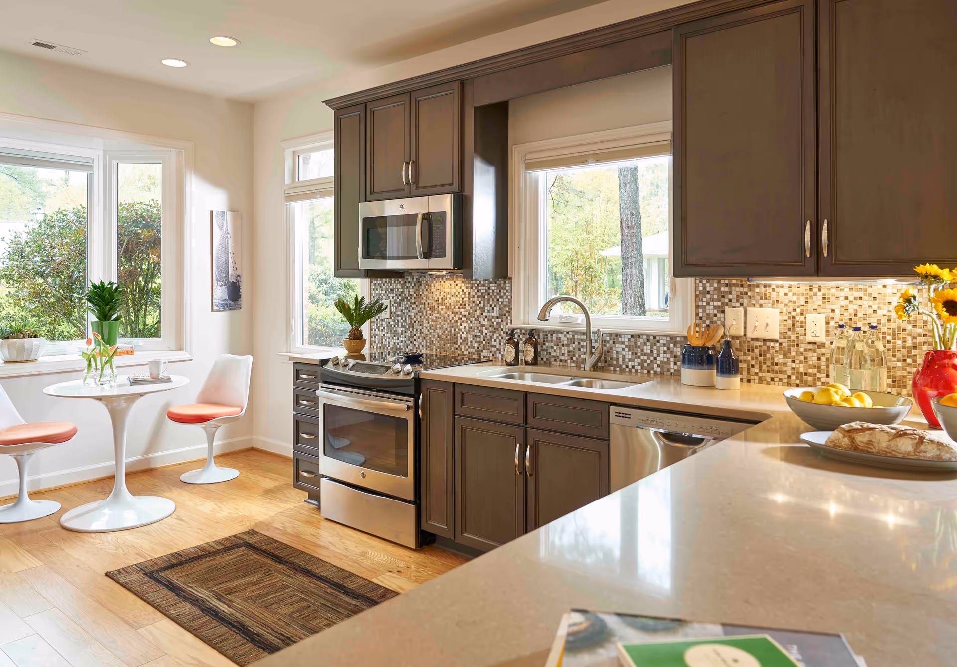 Bright modern kitchen with dark cabinets, stainless steel appliances, a double sink, mosaic backsplash and a small round breakfast table by windows.