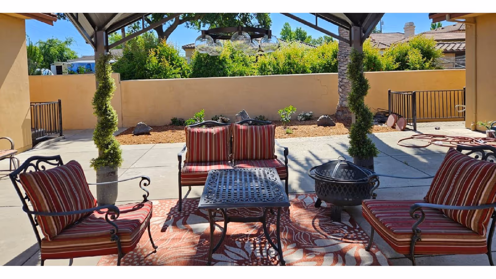 Outdoor patio area with a metal table, four cushioned chairs with red and white striped upholstery, a fire pit, and two potted spiral topiary plants under a shaded pergola. The background shows a beige wall, some greenery, and neighboring houses under a clear blue sky.