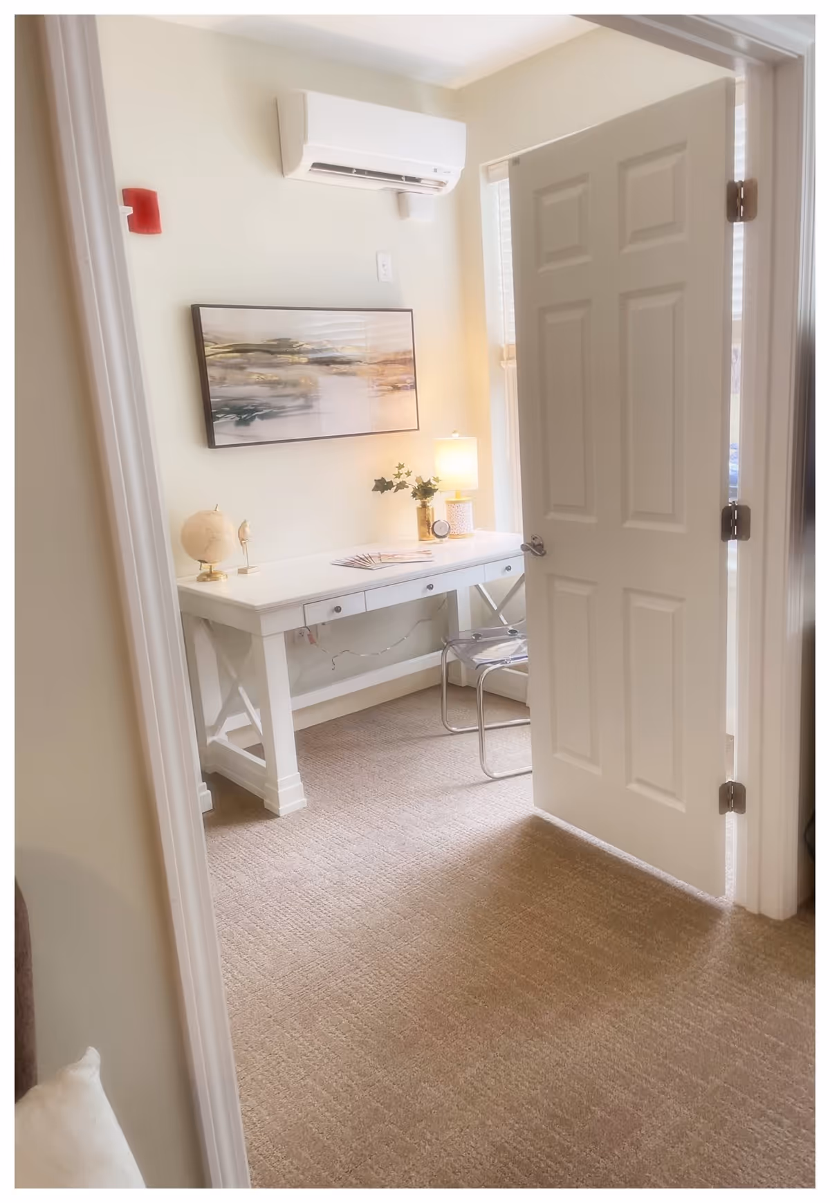 View through an open door into a small room with beige carpet and light-colored walls. Inside the room is a white desk with three drawers, a small globe, a decorative bird, a lamp, a small clock, and some greenery in a vase. Above the desk hangs a framed abstract landscape painting. A transparent chair is placed in front of the desk. A white air conditioning unit is mounted on the wall above the painting.