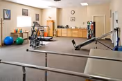 Exercise/rehab room with parallel bars, exercise equipment, therapy table, balance balls, and cabinets in a senior care facility.