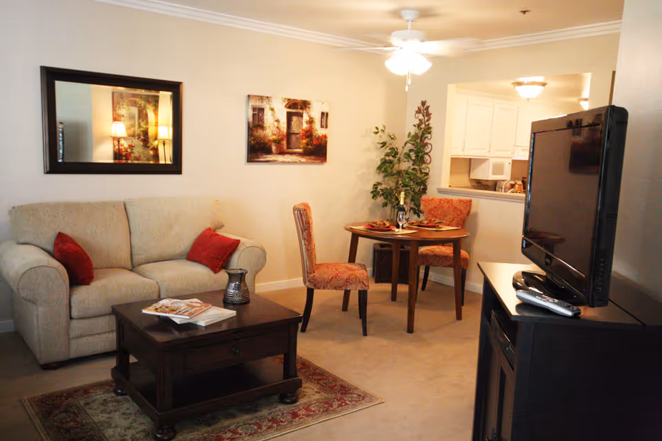 Cozy living room with a beige sofa, coffee table, TV and a small dining table near a kitchen pass-through.