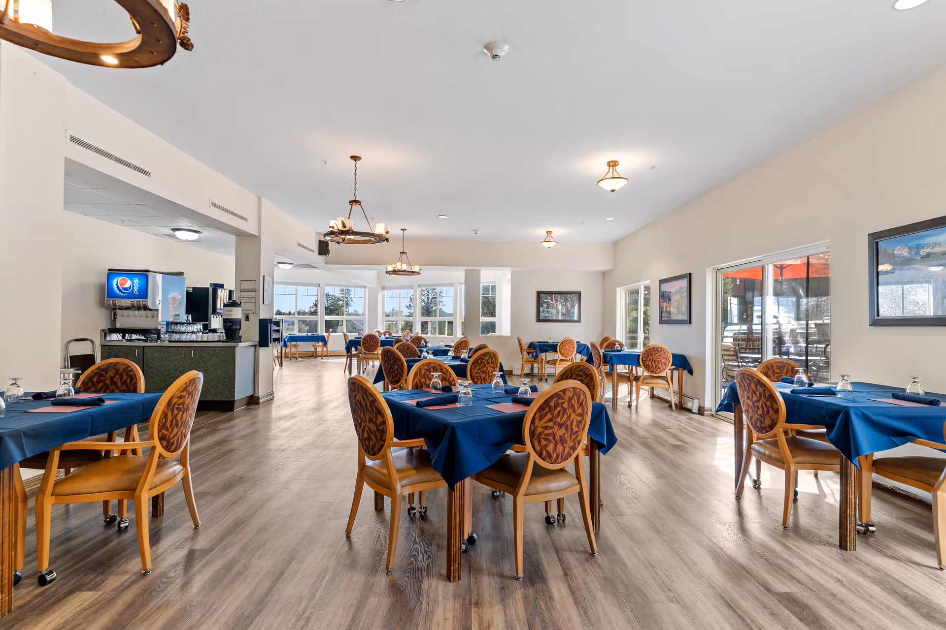 A spacious dining room in an assisted living facility with multiple tables covered in blue tablecloths, each set with glasses and napkins. The room features wooden chairs with patterned upholstery, large windows allowing natural light, and a beverage station with a soda dispenser in the background. The floor is wood, and there are framed pictures on the walls.
