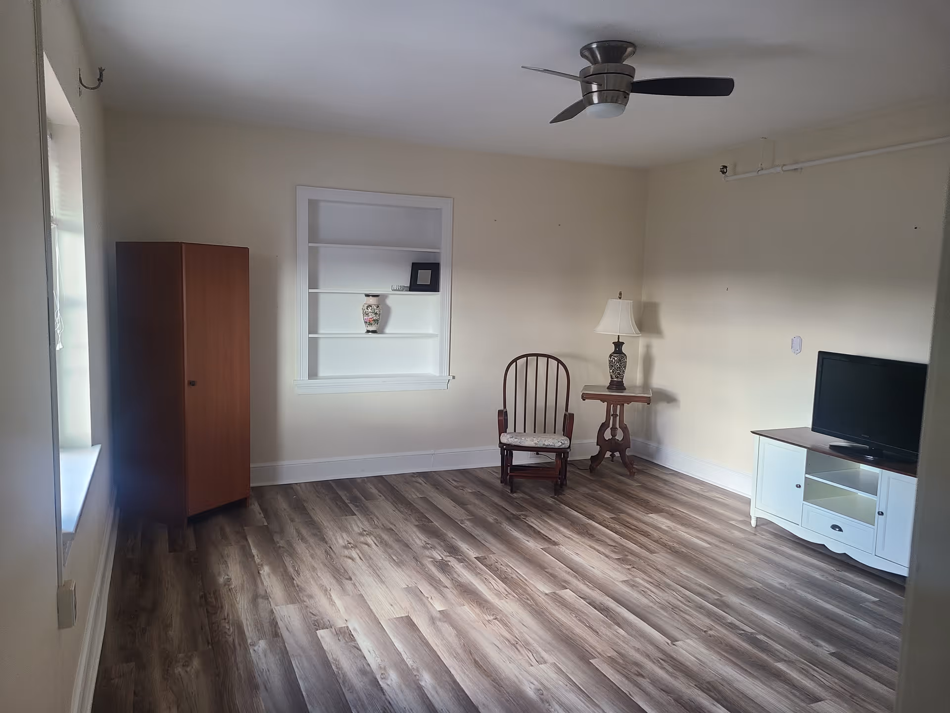 A sparsely furnished room with light-colored walls and wood flooring. The room contains a wooden cabinet near a window, a built-in white shelf with a decorative vase and a small framed item, a wooden rocking chair with a cushioned seat, a small wooden side table with a lamp, and a white TV stand with a flat-screen television. A ceiling fan with dark blades is mounted on the ceiling.