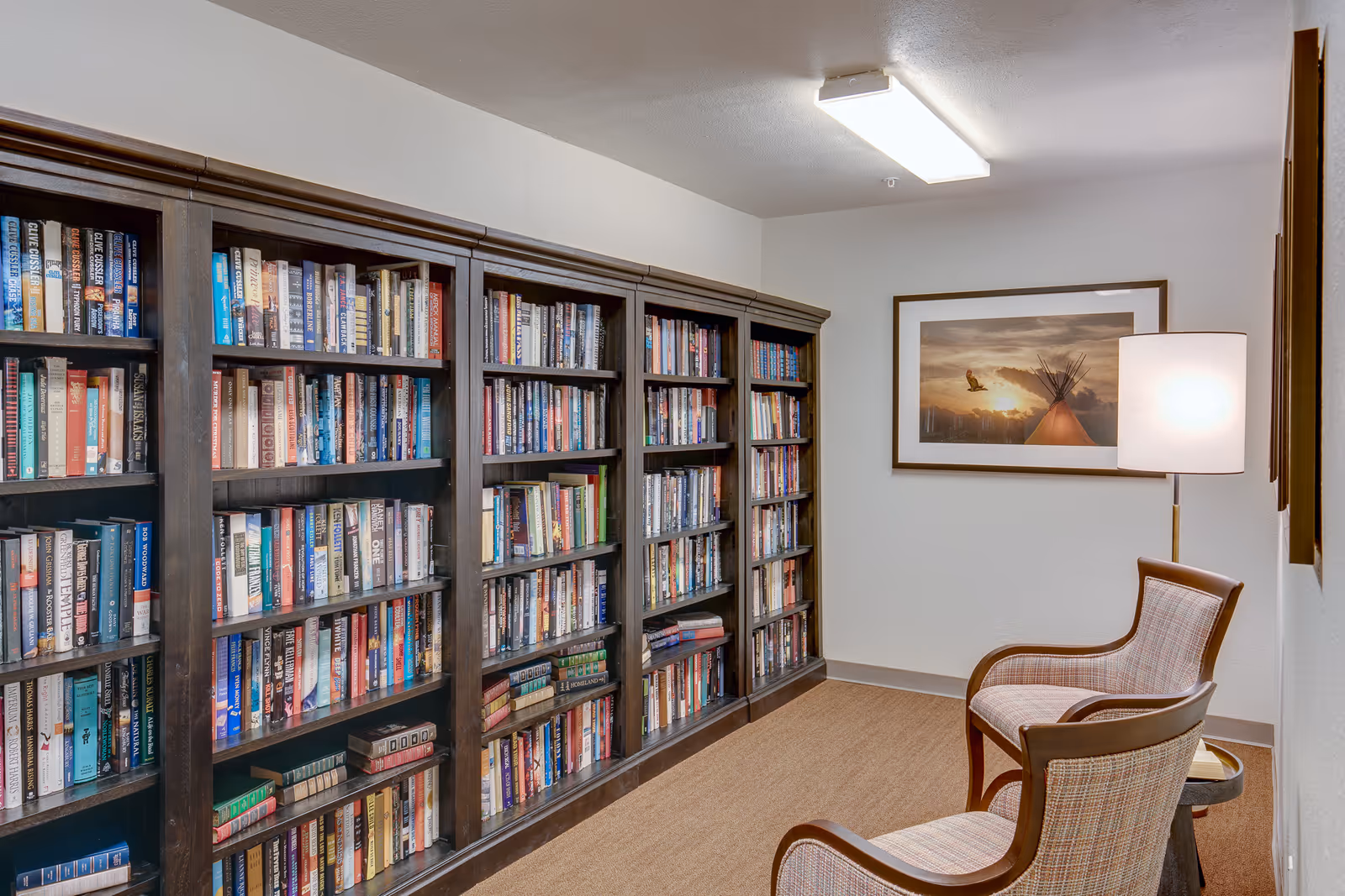 A cozy reading nook in a senior living facility featuring a large dark wood bookshelf filled with books, two upholstered armchairs, a floor lamp, and a framed picture of a sunset with a bird and a teepee on the wall.