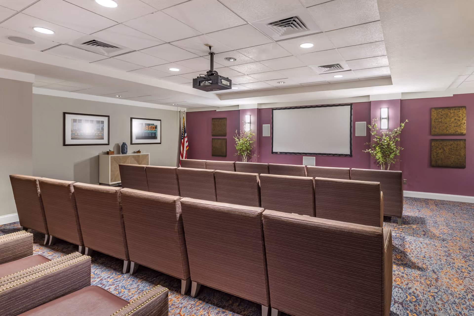 A small theater room with rows of brown cushioned chairs facing a large projector screen mounted on a purple accent wall. The room has a patterned carpet, framed pictures on the walls, two potted plants on either side of the screen, and an American flag in the corner. Ceiling lights and a ceiling-mounted projector are also visible.