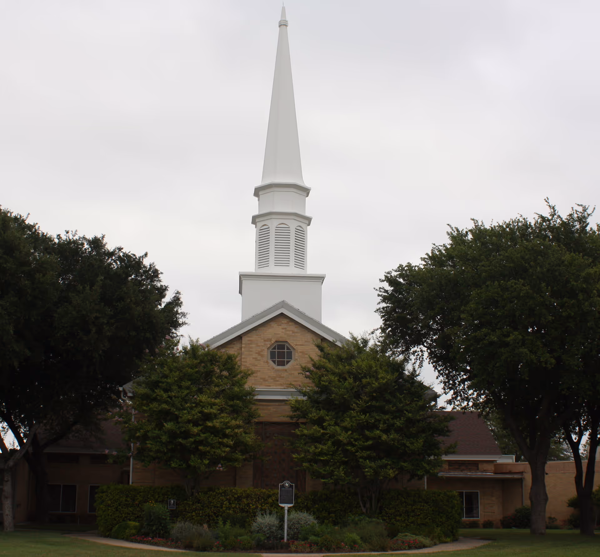 Exterior view of a building with a tall white steeple, surrounded by trees and greenery under a cloudy sky.