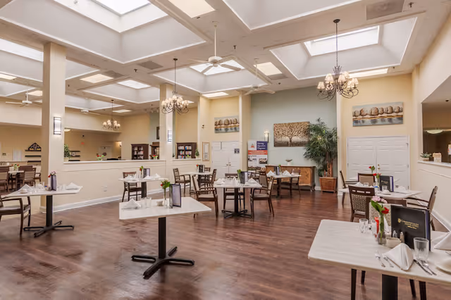 A spacious dining room with several tables set with white napkins, glasses, and small flower vases. The room features wooden flooring, cream-colored walls, chandeliers, ceiling fans, and skylights allowing natural light. There are paintings and plants decorating the walls and corners.