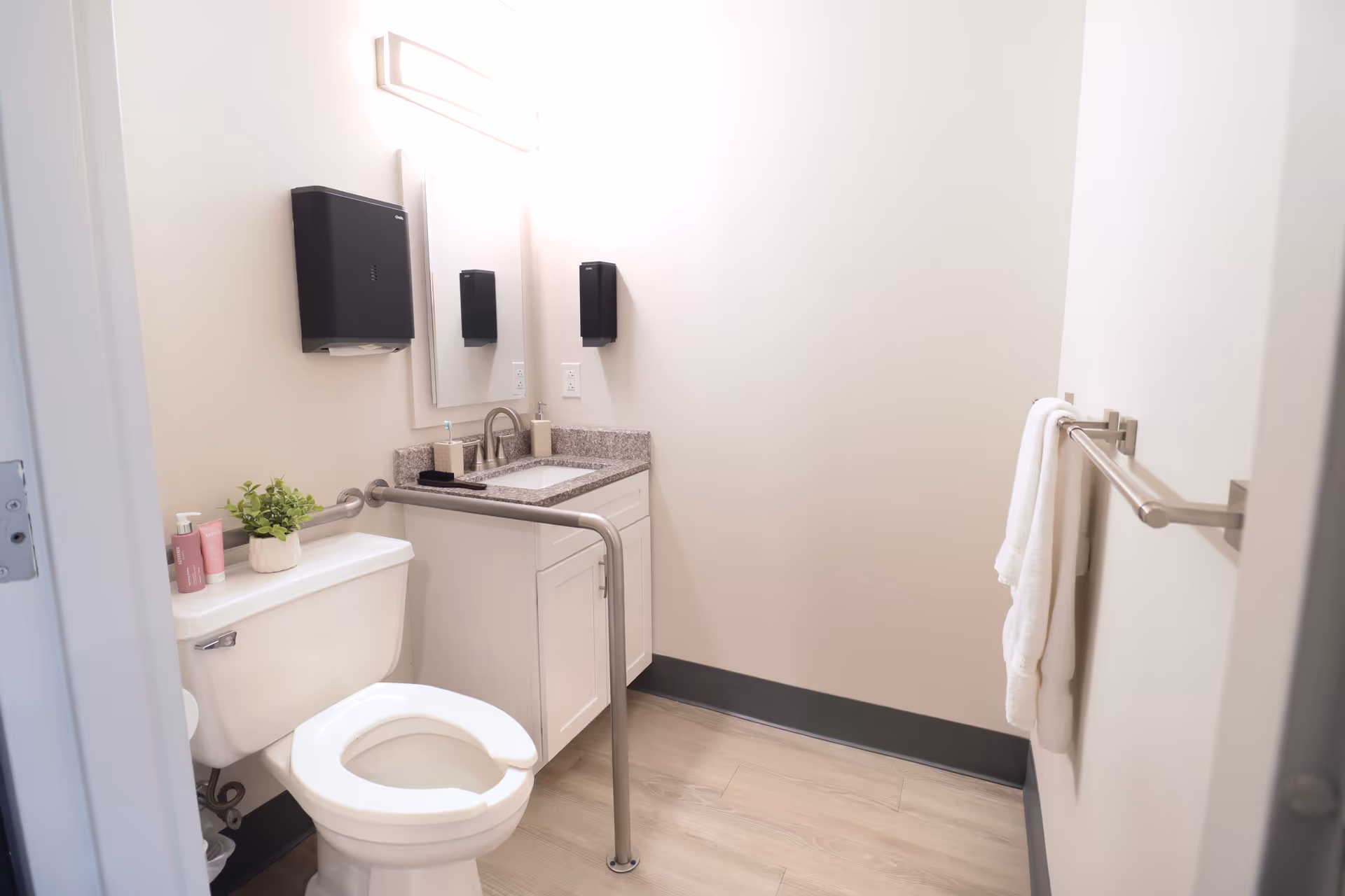 A clean and bright bathroom with a white toilet, a white vanity with a granite countertop, a mirror above the sink, a black paper towel dispenser, a soap dispenser, a small potted plant on the toilet tank, and a towel rack with white towels on the right wall. The bathroom has light-colored walls and wood-like flooring.
