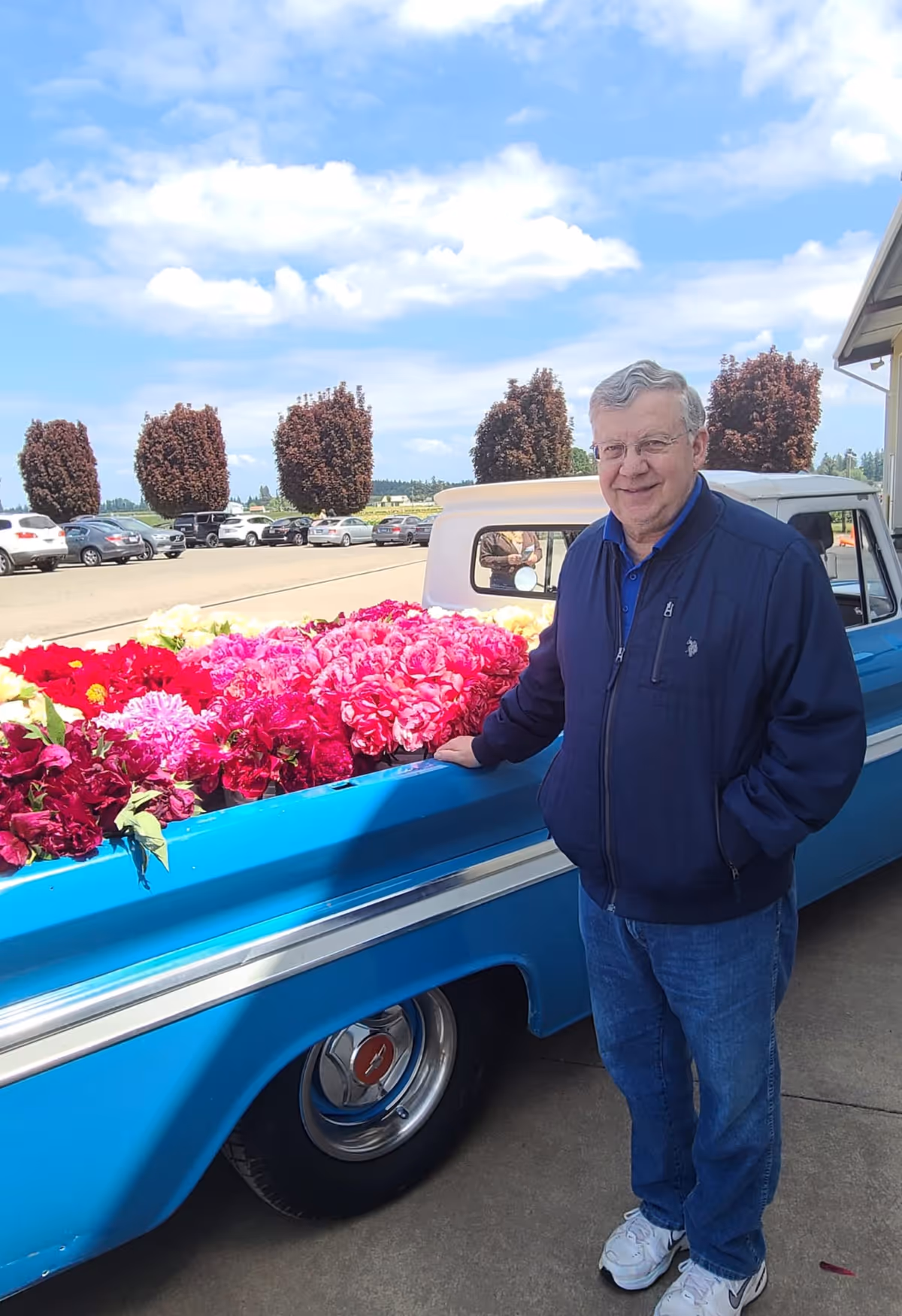 An elderly man wearing a navy blue jacket, blue jeans, and white sneakers stands next to a blue vintage pickup truck filled with vibrant red, pink, and white flowers. The scene is outdoors on a sunny day with a partly cloudy sky, parked cars, and neatly trimmed trees in the background.