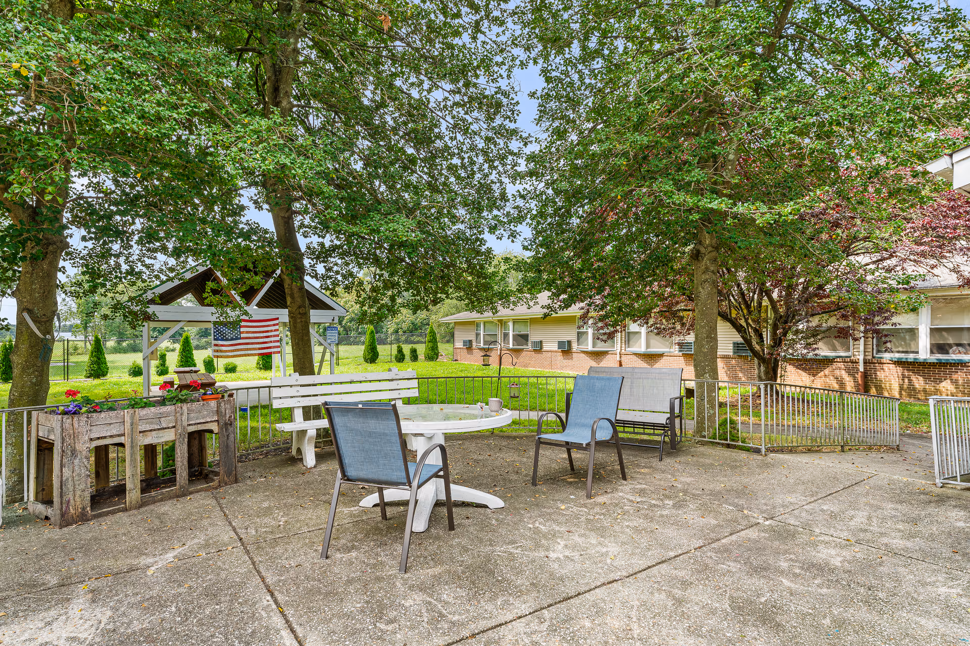 Outdoor patio area with a white round table surrounded by two blue chairs and a white bench. There are large trees providing shade, a wooden planter box with flowers, and an American flag hanging from a small pavilion. A brick building with multiple windows is visible in the background.