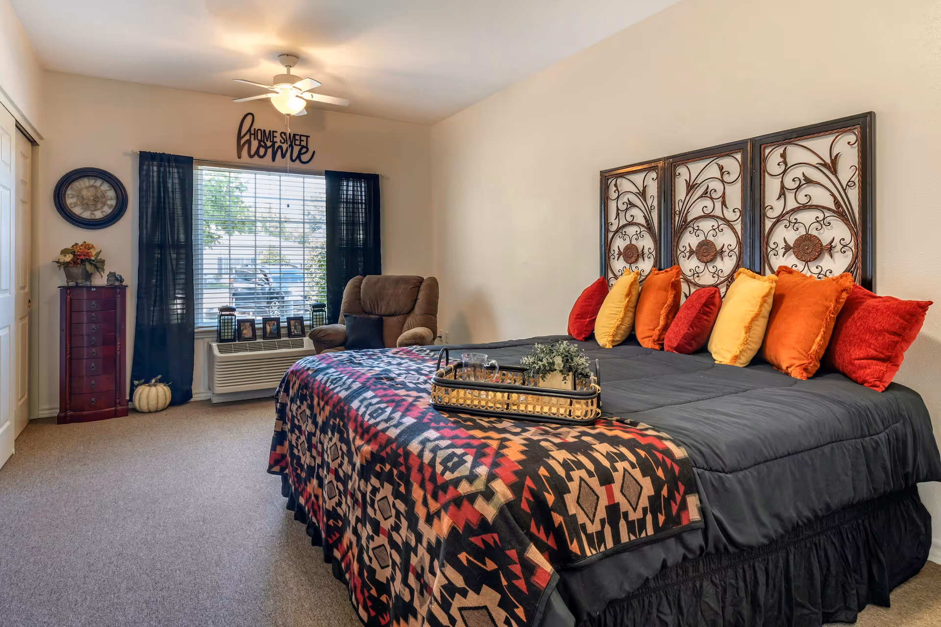A cozy bedroom with a large bed covered in a black comforter and a colorful patterned blanket. The bed has multiple decorative pillows in red, orange, and yellow. Above the bed are three ornate metal wall hangings. To the left, there is a window with black curtains, a brown recliner chair, and a small dresser with a clock and flowers on top. A ceiling fan with a light is on the ceiling, and a 'Home Sweet Home' sign is above the window.